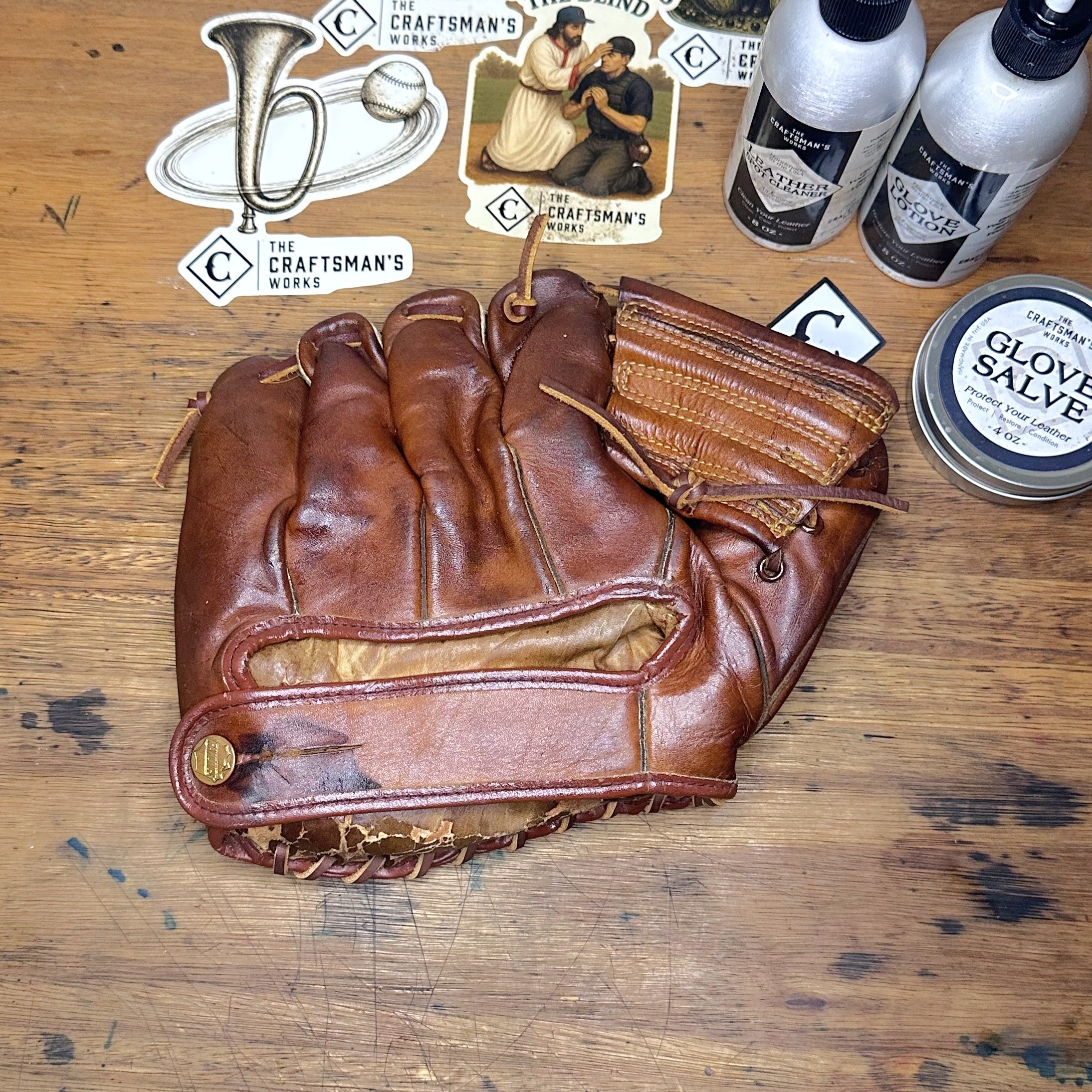 A brown leather baseball glove is laid flat on a wooden surface, surrounded by Craftsmen's Wipes stickers, two bottles of liquid cleaner, and a tin of clove salve.