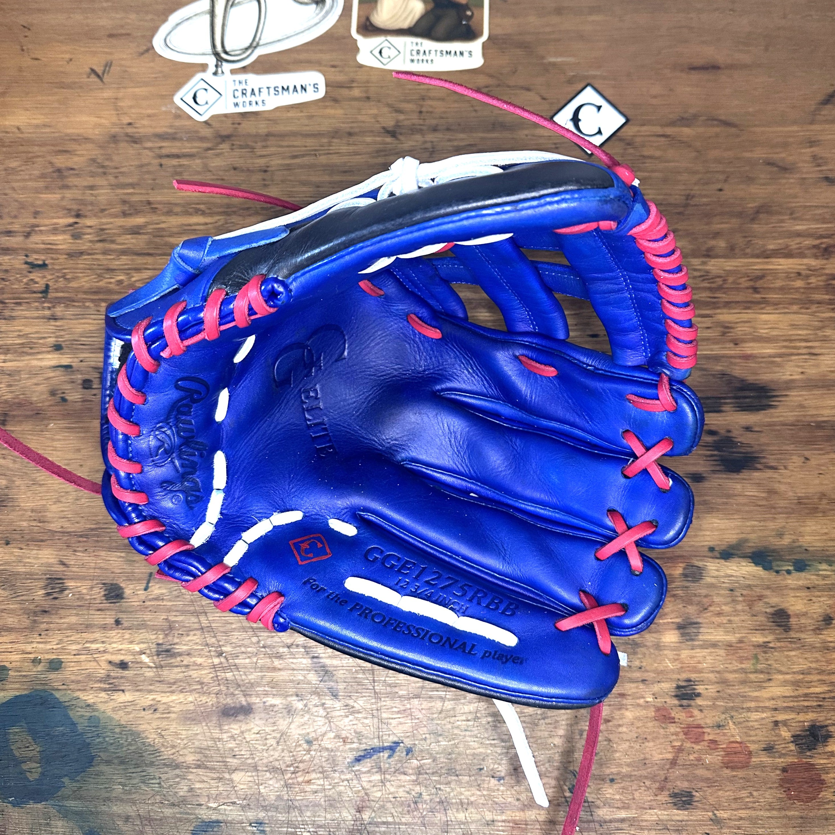 A blue leather baseball glove with bright pink laces and white stitching, laid flat on a rustic wooden table. The glove features the Cactus Jack brand logo and 'G.