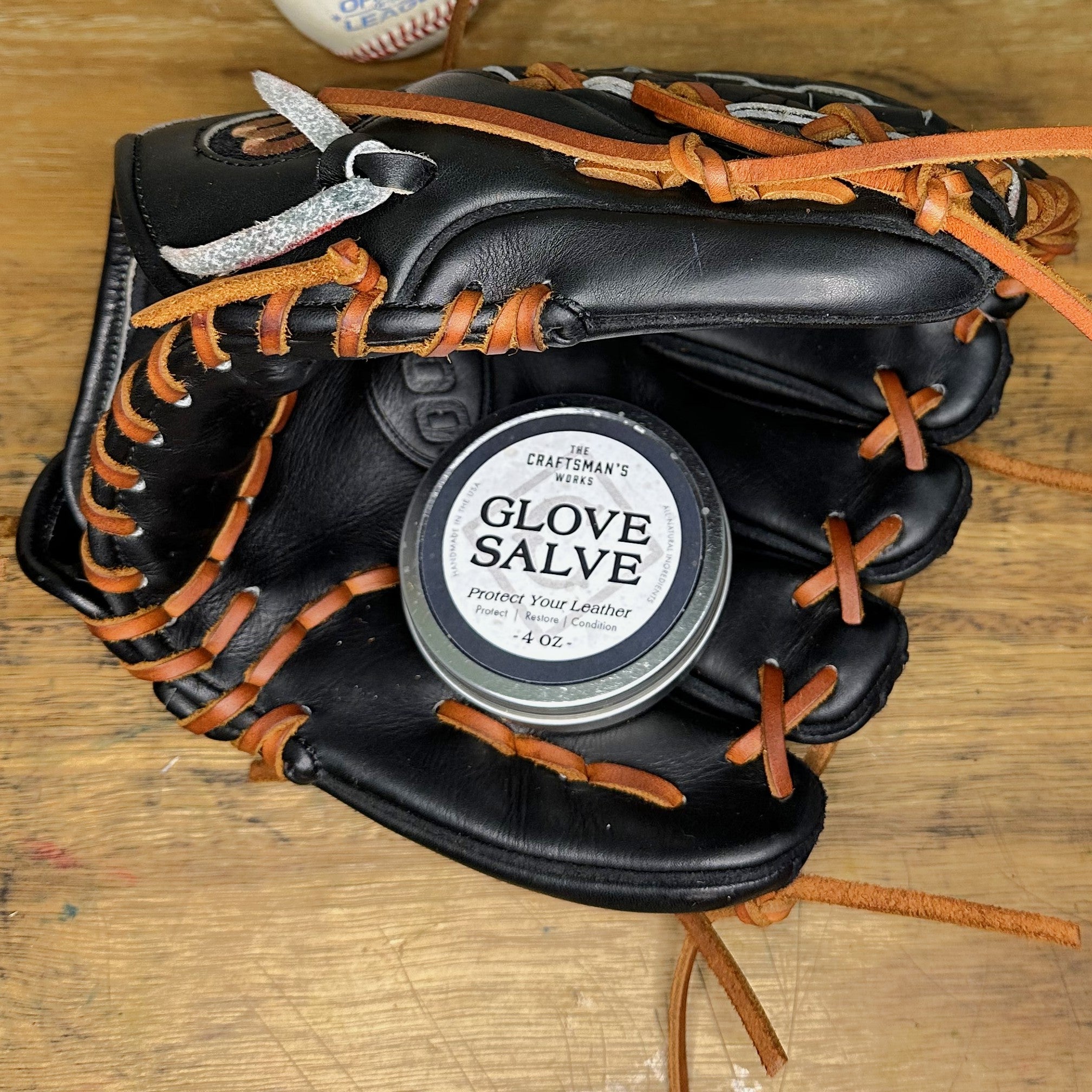 A tin container labeled 'GLOVE SALVE' placed on a leather glove with laces, indicative of its use for conditioning and protecting leather products.