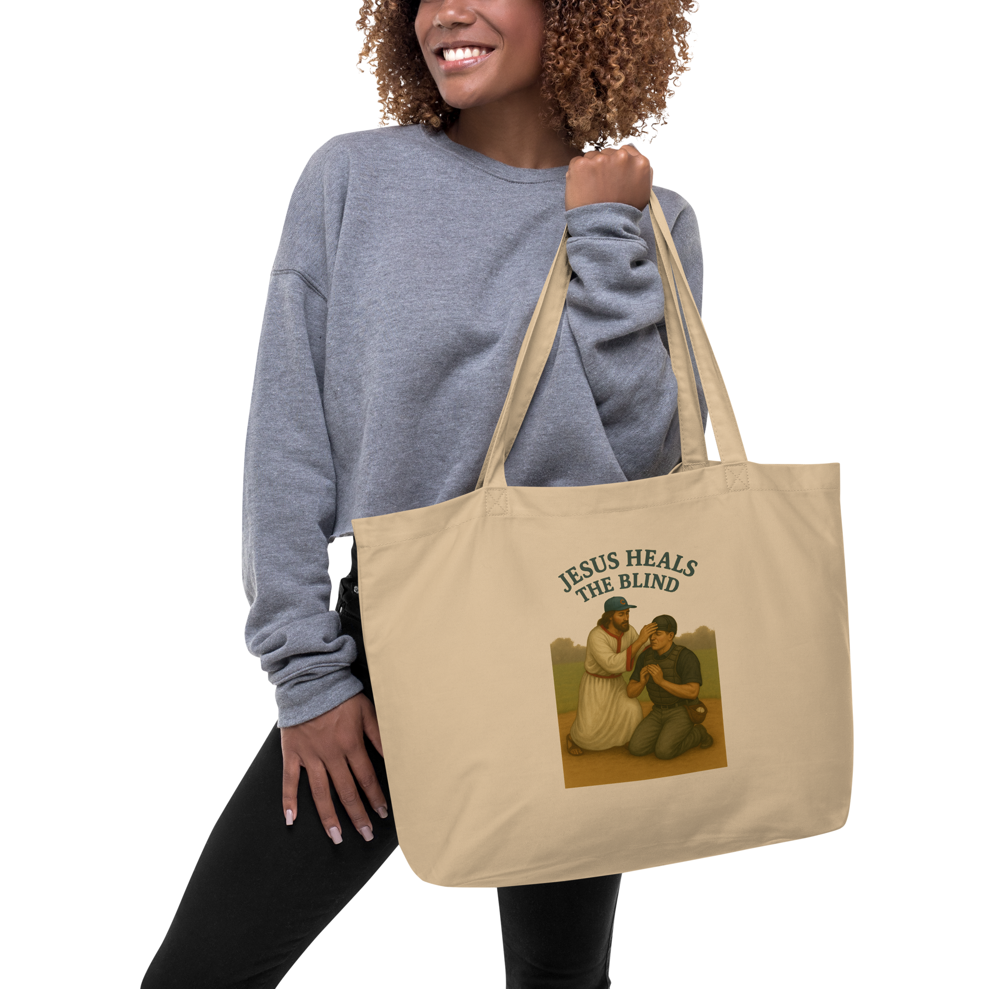 A woman wearing a gray sweatshirt and black pants carries a beige canvas tote bag with a printed illustration and the text 'JESUS HEALS THE BLIND' on a plain white background.