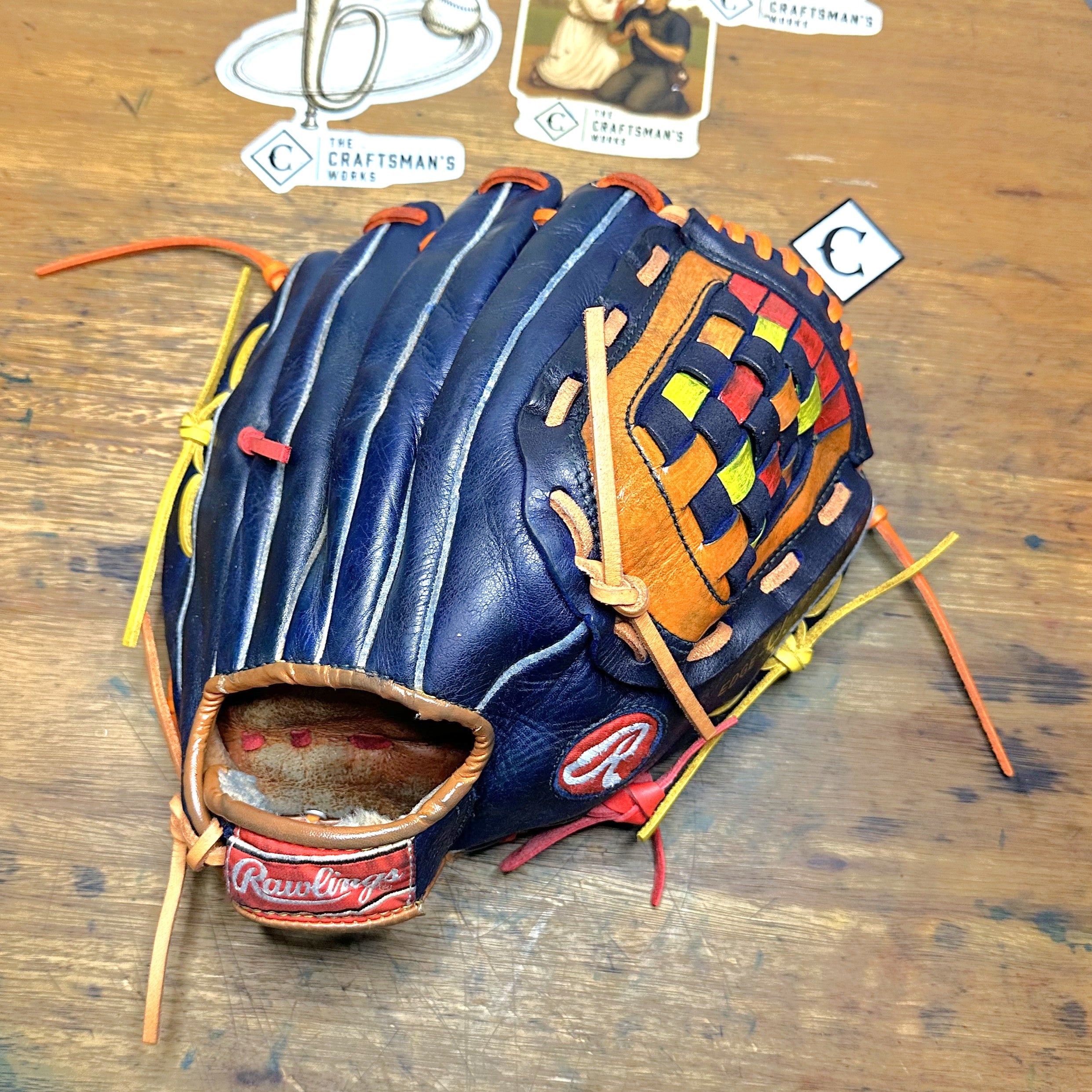 A Rawlings baseball glove with a navy blue leather back and a multicolor woven pattern in yellow, red, and green on the palm, displayed on a wooden tabletop with brand cards in the background.