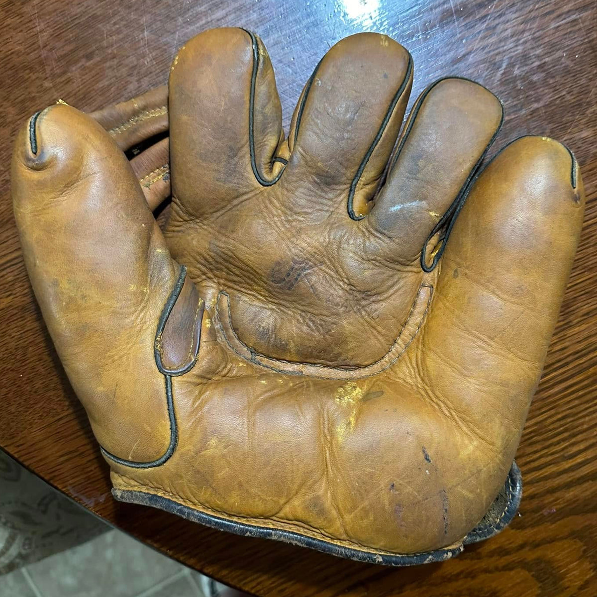 A single tan leather baseball glove laid flat on a wooden table, photographed from above. A person's bare feet are visible at the bottom edge, and part of a room floor is visible in the lower left.