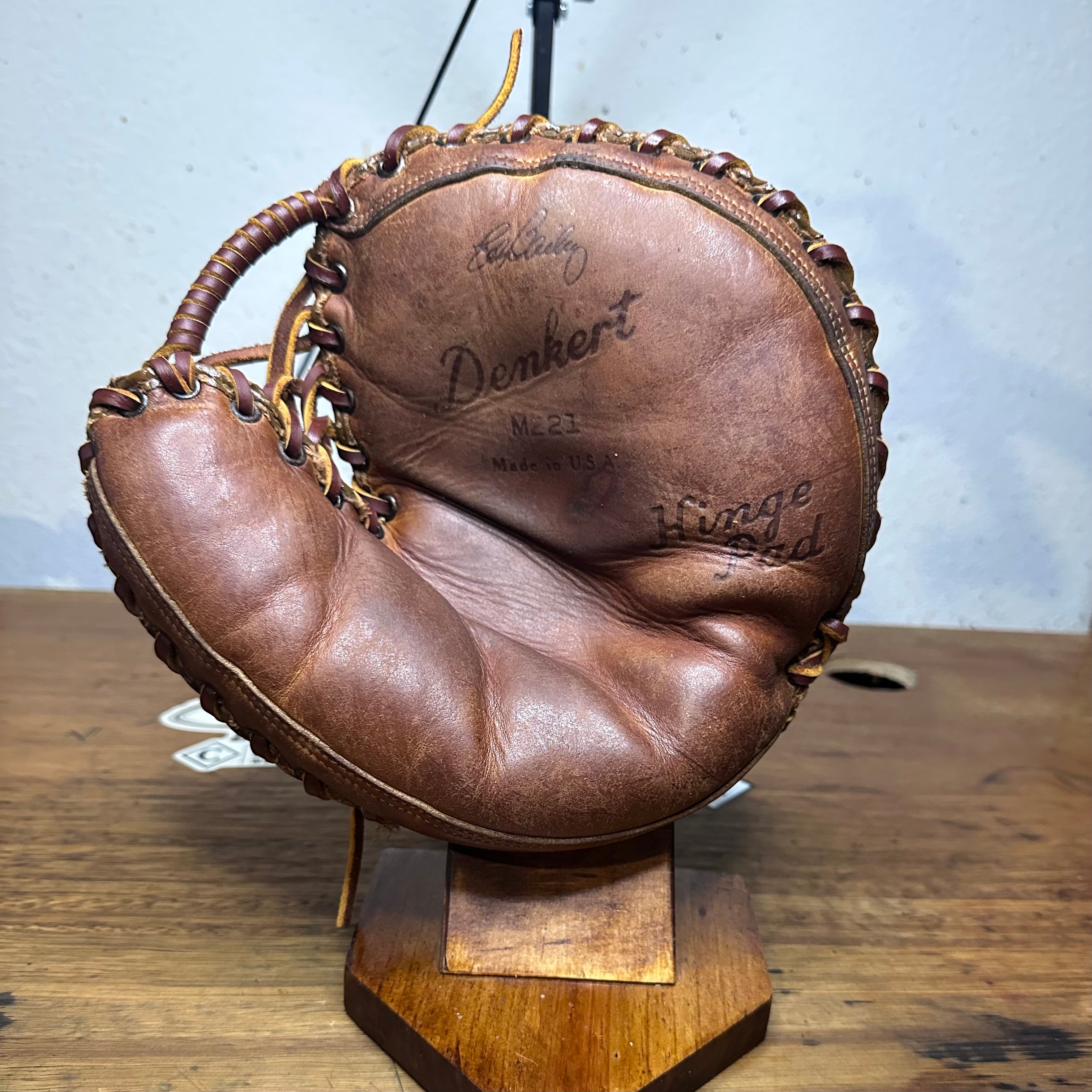 A vintage brown leather baseball glove displayed on a wooden stand, featuring embossed text including 'Denkert' and 'Mays'.