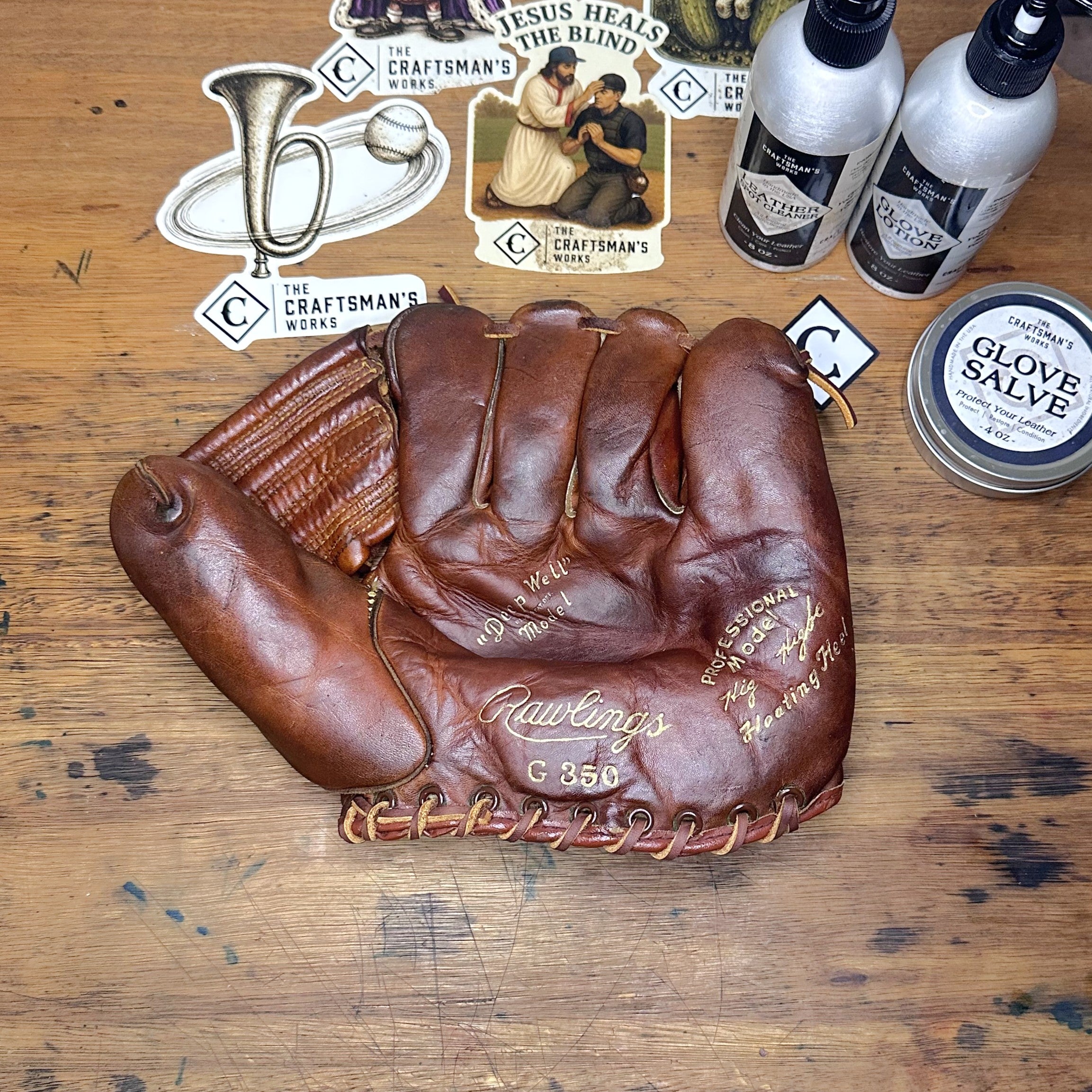 A brown leather baseball glove is laid flat on a wooden surface, surrounded by vintage-style stickers, two white bottles, and a round tin labeled glove salve. The glove has visible branding and cursive script on the palm.