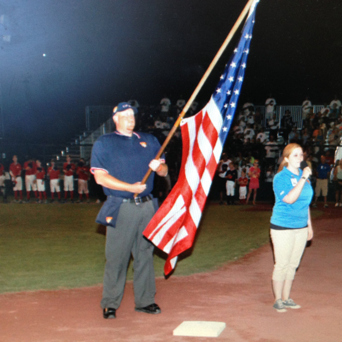 An umpire holding an american flag during the national anthem at a game at Cooperstown Dreams park