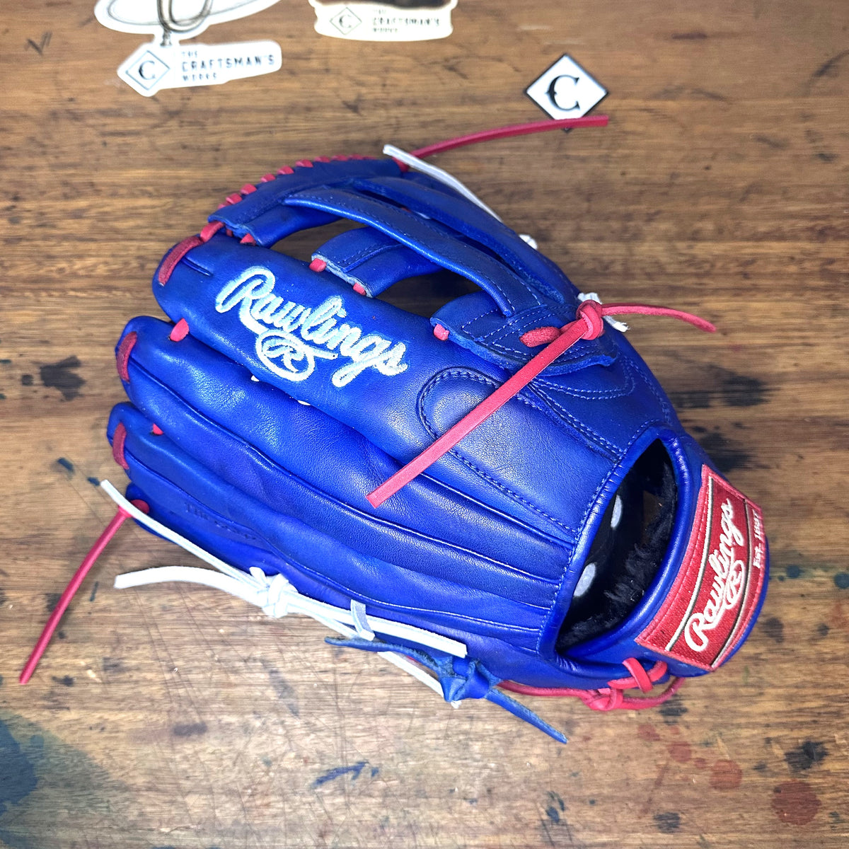 A blue Rawlings baseball glove with red and white laces and stitching, placed on a wooden tabletop. Rawlings branding is visible on the glove and attached tags are also present.