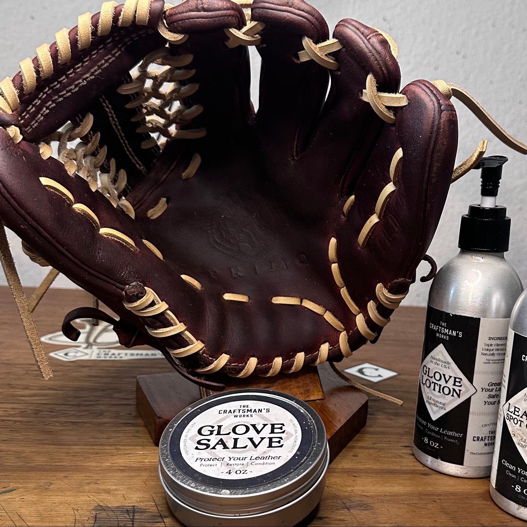 A brown leather baseball glove with cream lacing displayed on a wooden stand on a tabletop, with a tin of glove salve in front and two bottles of leather care products to the right.