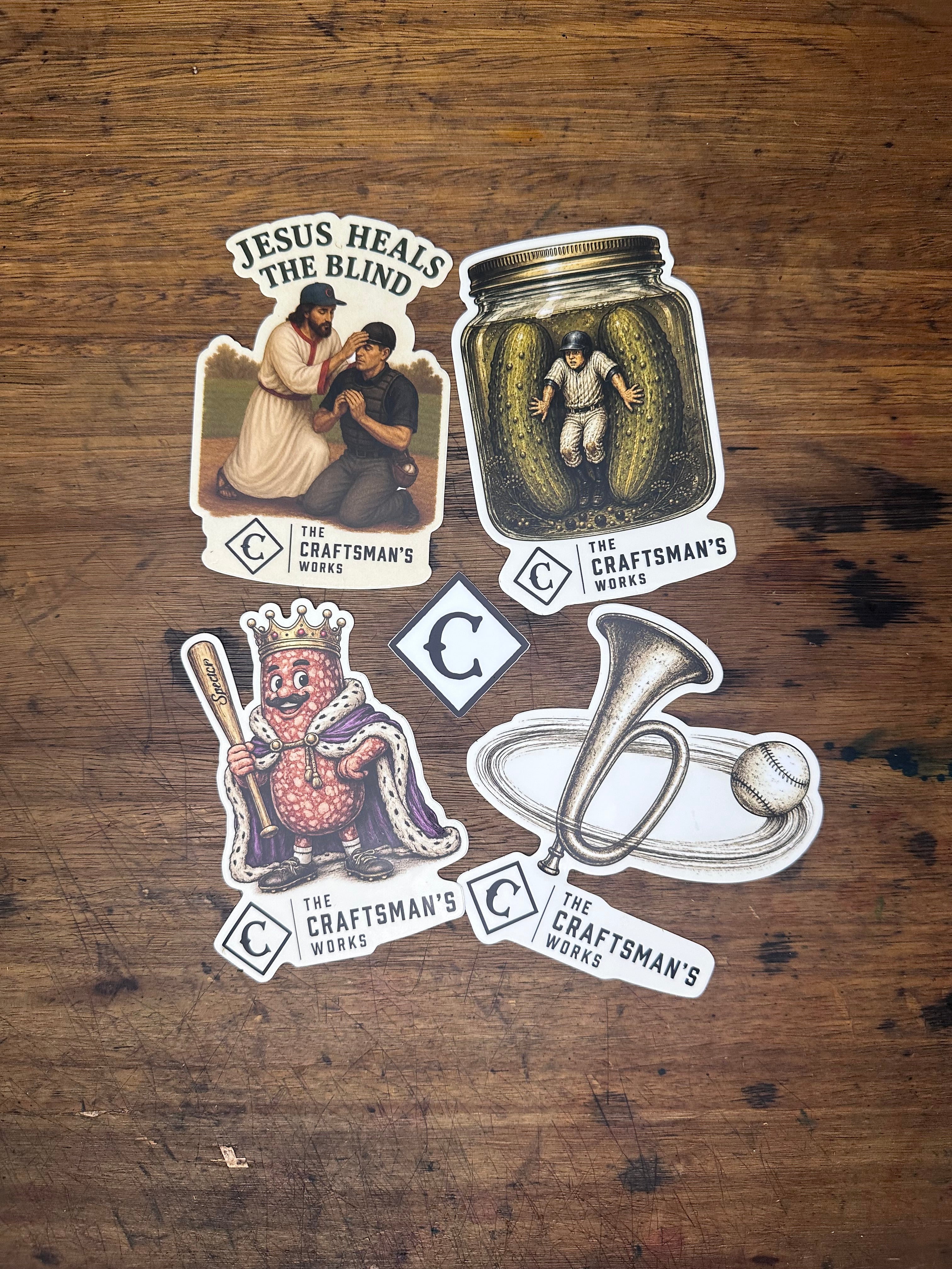 Five die-cut stickers from The Craftsman's World arranged on a rustic wooden surface. The stickers feature vintage-style illustrations, including a pickle jar, a trumpet, a couple embracing, a crowned figure, and a central diamond logo.