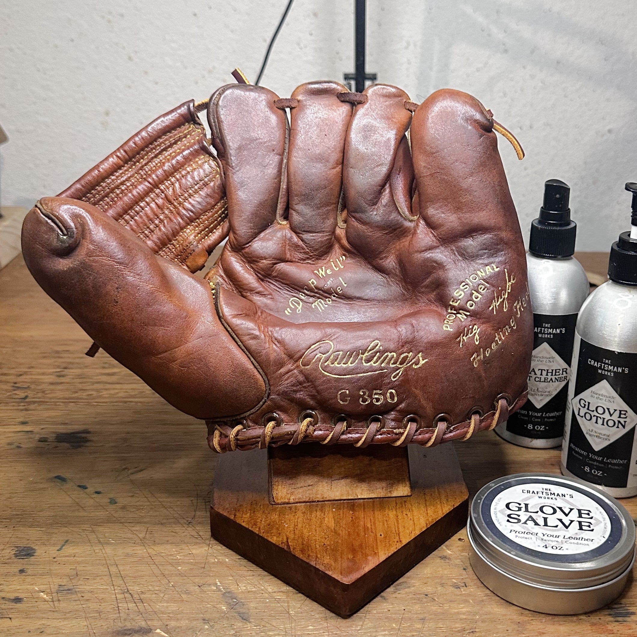 A brown leather baseball glove with visible signatures and 'Rawlings G 350' branding, displayed on a wooden stand on a wooden table. A tin of Glove Salve.