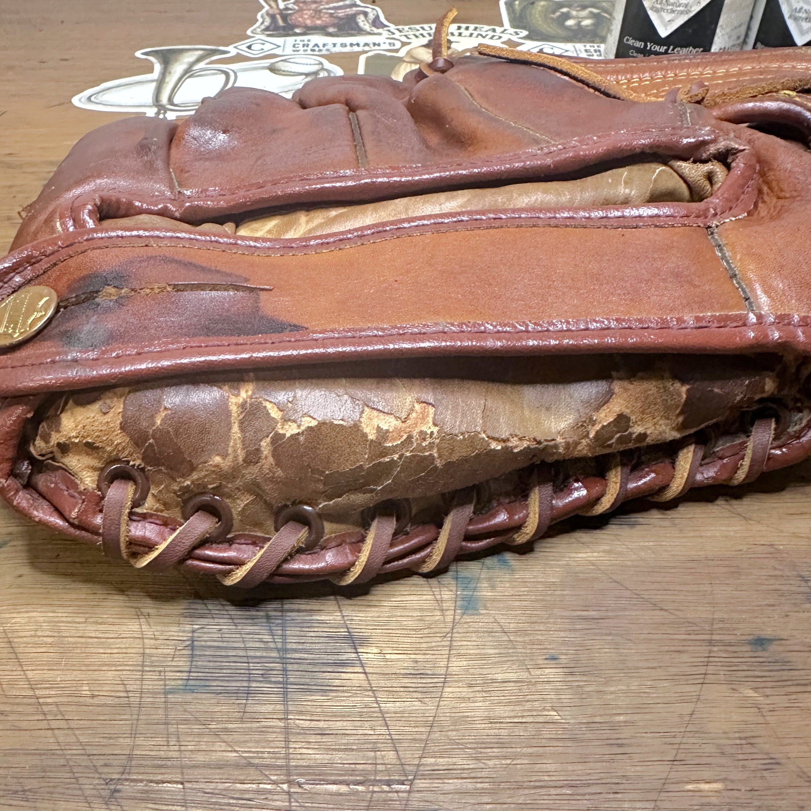 A close-up of a worn brown leather baseball glove resting on a scratched wooden tabletop. Damage on the inside of the glove.