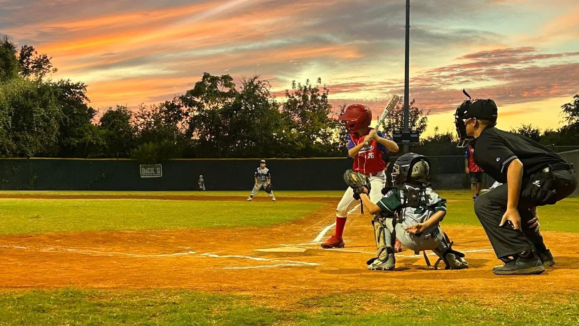A baseball game scene at sunset with a batter in a red helmet standing at home plate, a catcher in protective gear crouching, and an umpire in black observing. The field is green and brown dirt, with trees and a bright stadium light against a dramatic orange and blue sky.