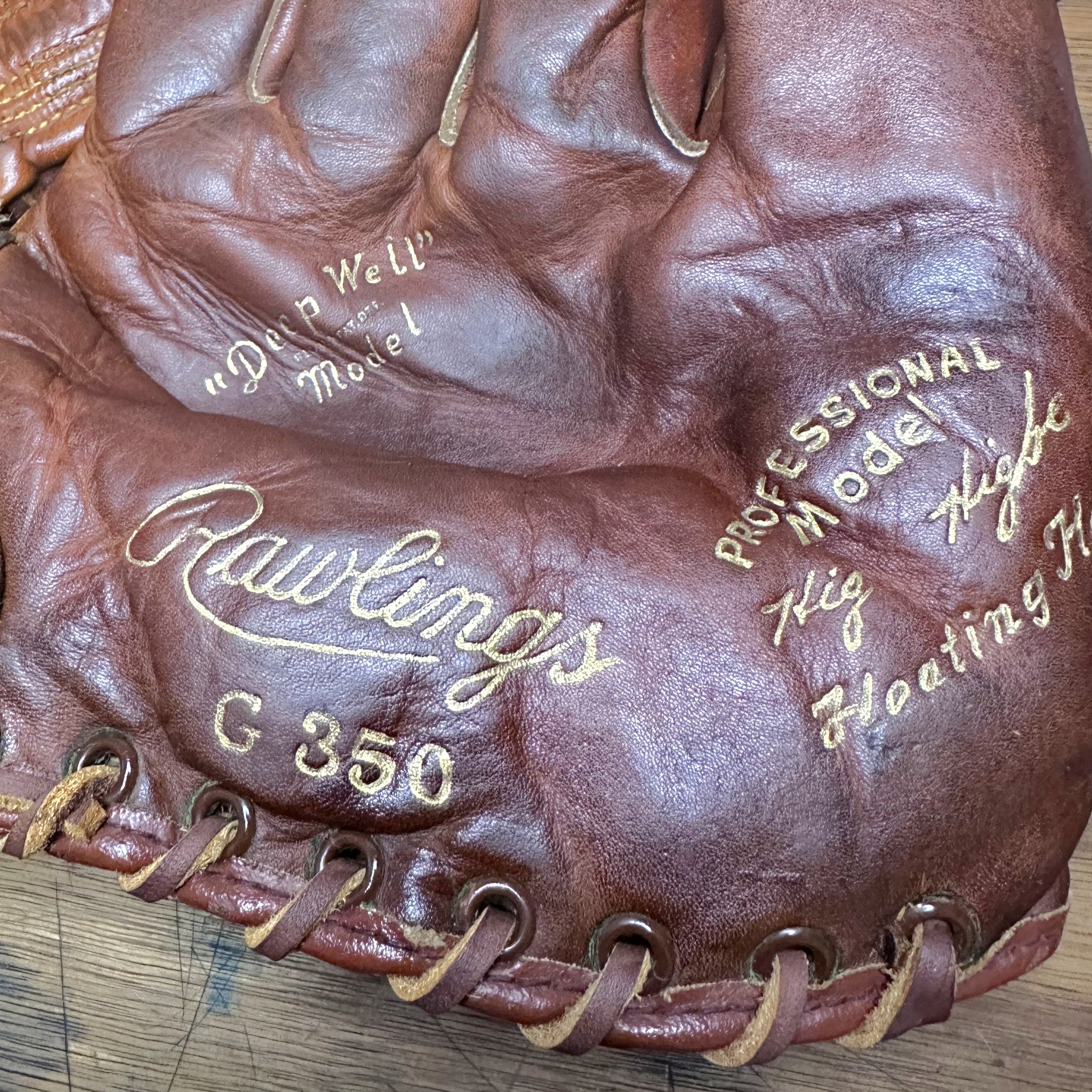 Close-up of a brown leather baseball glove showing the Rawlings G 350 branding and gold-embossed text on the palm, with visible laces and a wooden surface underneath.