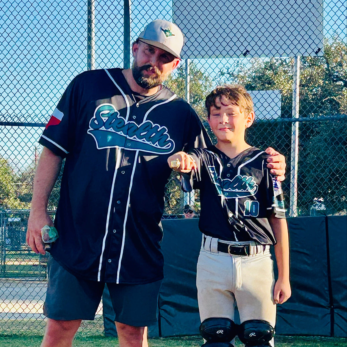 A man and a young boy wearing matching baseball jerseys with 'Sparta' on the front, standing together on a baseball field. The man wears dark shorts and a cap, and the boy wears gray pants and black shin guards.